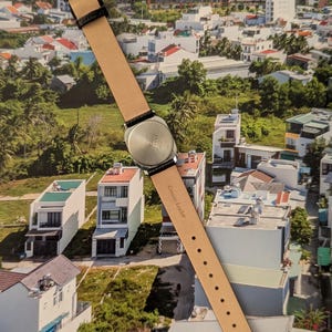 May include: A wristwatch with a silver-colored case and a light brown leather strap. The strap is attached to the watch with black leather accents. The watch is displayed against a backdrop of a cityscape.