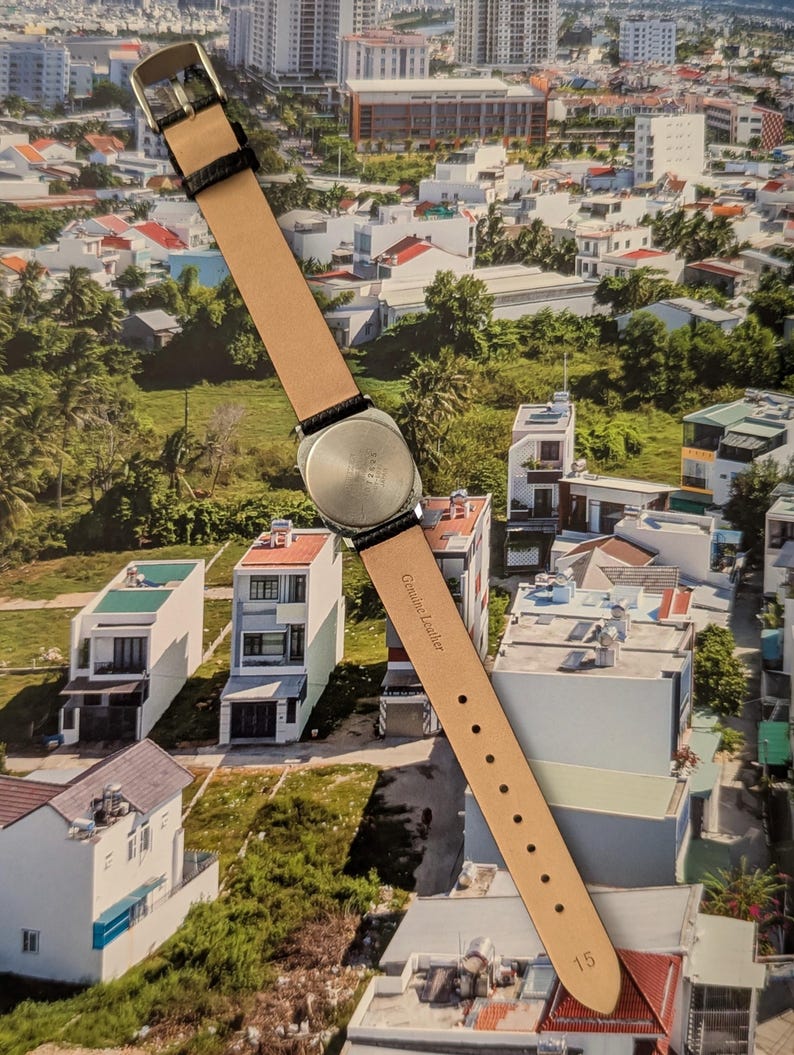 May include: A wristwatch with a silver-colored case and a light brown leather strap. The strap is attached to the watch with black leather accents. The watch is displayed against a backdrop of a cityscape.