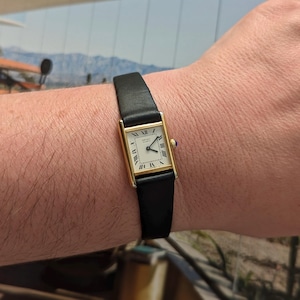 May include: A gold rectangular wristwatch with a white face and black Roman numerals. The watch has a black leather strap and a small blue gem on the side. The watch is approximately 2.5 cm wide.