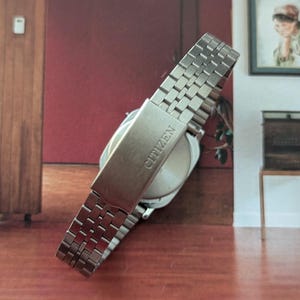 May include: A silver Citizen wristwatch with a stainless steel band. The watch band is composed of small, rectangular links. The clasp is visible and has the brand name "Citizen" engraved on it. The watch face is round and silver.