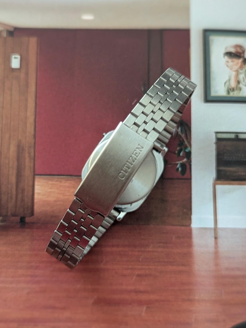 May include: A silver Citizen wristwatch with a stainless steel band. The watch band is composed of small, rectangular links. The clasp is visible and has the brand name "Citizen" engraved on it. The watch face is round and silver.