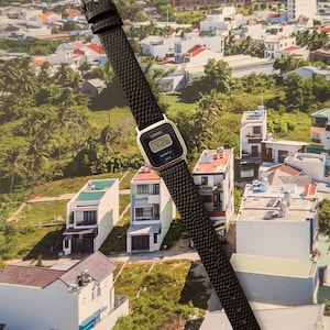 May include: A Citizen digital watch with a black textured leather band and a silver-tone buckle. The watch face displays the time, 4:26, in a digital format. The watch is set against a backdrop of a cityscape.