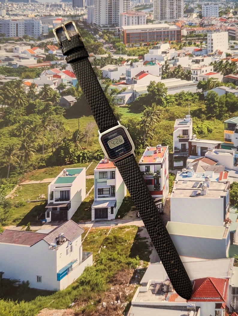 May include: A Citizen digital watch with a black textured leather band and a silver-tone buckle. The watch face displays the time, 4:26, in a digital format. The watch is set against a backdrop of a cityscape.