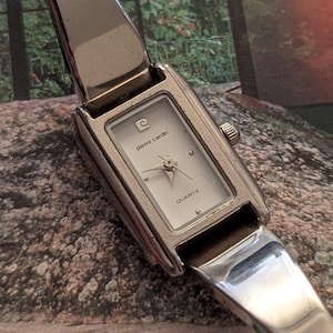 Może przedstawiać: Srebrny zegarek kwarcowy Pierre Cardin z prostokątną tarczą. Biała tarcza z logo Pierre Cardin i napisem "QUARTZ". Pasek zegarka jest srebrny, z zawiasami.
