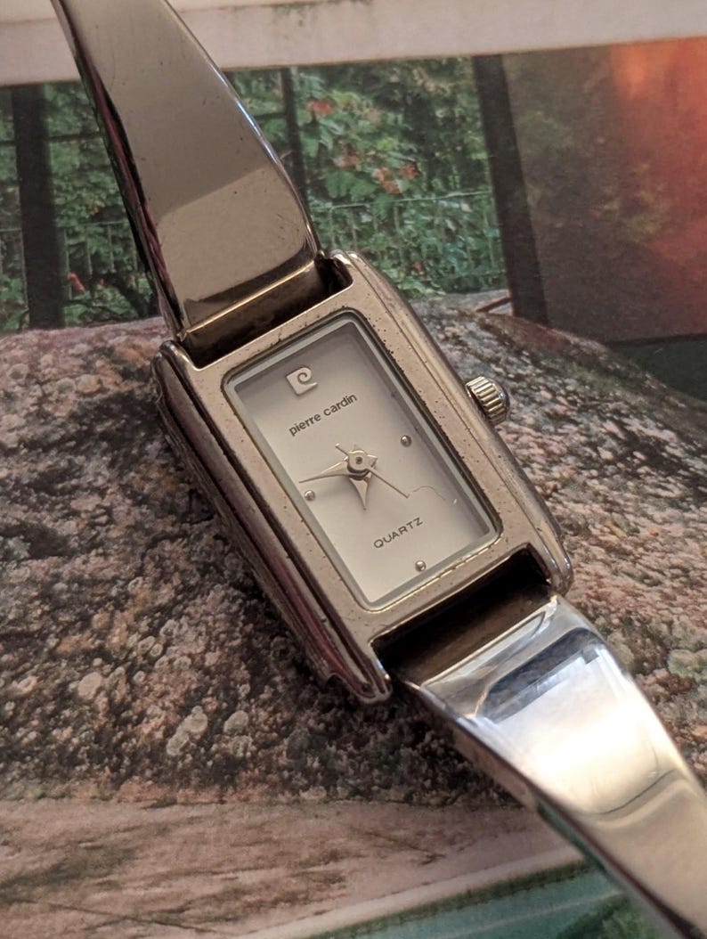 Może przedstawiać: Srebrny zegarek kwarcowy Pierre Cardin z prostokątną tarczą. Biała tarcza z logo Pierre Cardin i napisem "QUARTZ". Pasek zegarka jest srebrny, z zawiasami.