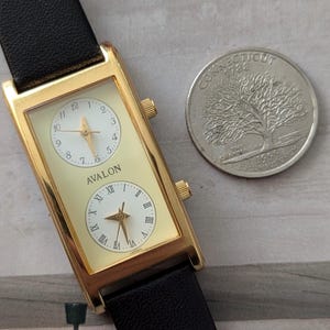 Puede incluir: Reloj rectangular dorado con dos esferas, correa de cuero negro y el nombre "AVALON" en la esfera. Una moneda de Connecticut est&aacute; junto al reloj. El reloj es de origen japon&eacute;s.