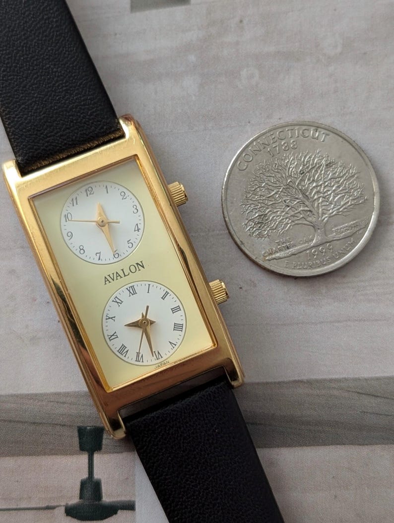 Puede incluir: Reloj rectangular dorado con dos esferas, correa de cuero negro y el nombre "AVALON" en la esfera. Una moneda de Connecticut est&aacute; junto al reloj. El reloj es de origen japon&eacute;s.