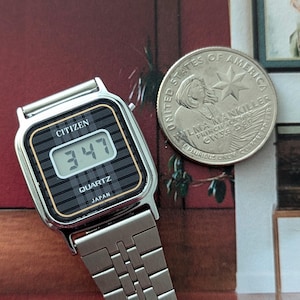 May include: A silver Citizen Quartz digital watch with a black and gold face displaying the time 3:47. The watch has a silver band and the text "JAPAN" is visible. A United States quarter coin is next to the watch.