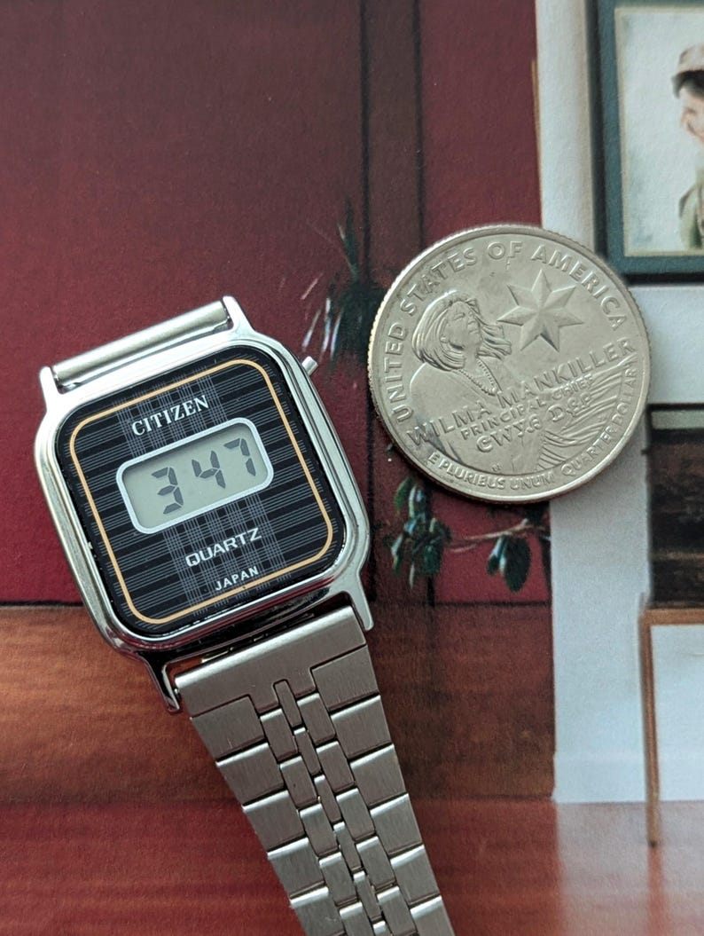 May include: A silver Citizen Quartz digital watch with a black and gold face displaying the time 3:47. The watch has a silver band and the text "JAPAN" is visible. A United States quarter coin is next to the watch.