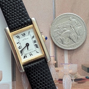 Peut inclure: Une montre rectangulaire dor&eacute;e avec un cadran blanc, des chiffres romains noirs et un bracelet en cuir noir. Le cadran de la montre indique "Le Grand" et "17 Jewels". Une pi&egrave;ce de monnaie en argent avec Theodore Roosevelt est &agrave; c&ocirc;t&eacute; de la montre.