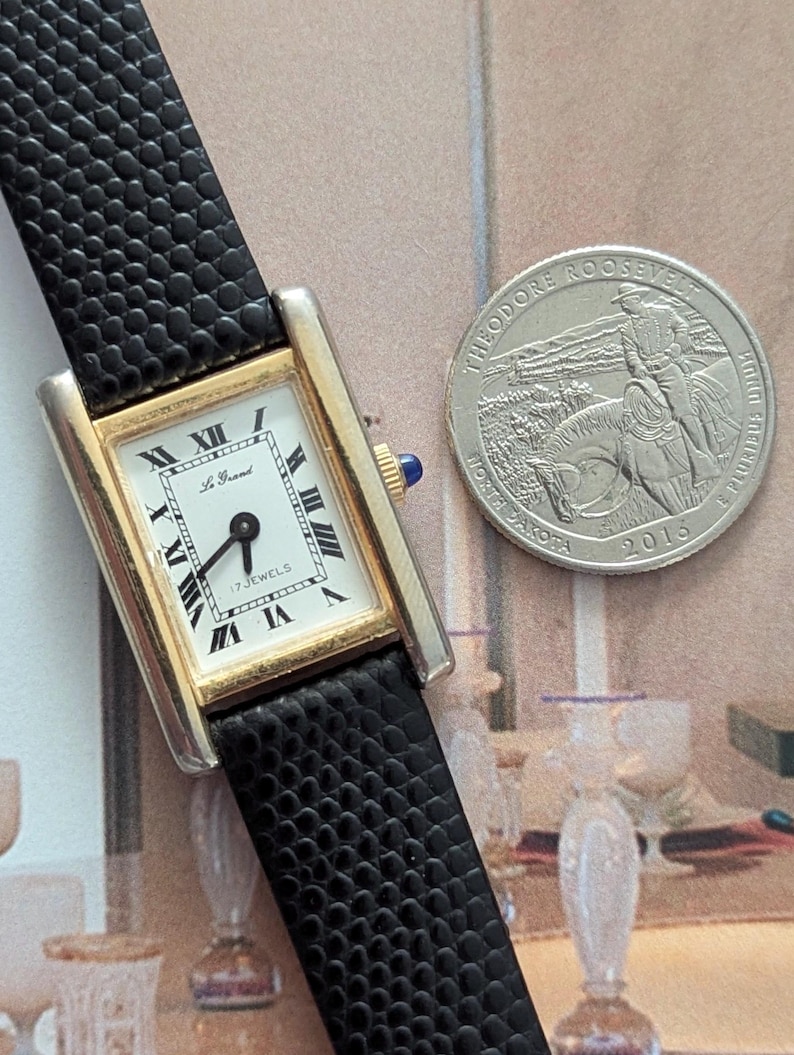 Peut inclure: Une montre rectangulaire dor&eacute;e avec un cadran blanc, des chiffres romains noirs et un bracelet en cuir noir. Le cadran de la montre indique "Le Grand" et "17 Jewels". Une pi&egrave;ce de monnaie en argent avec Theodore Roosevelt est &agrave; c&ocirc;t&eacute; de la montre.
