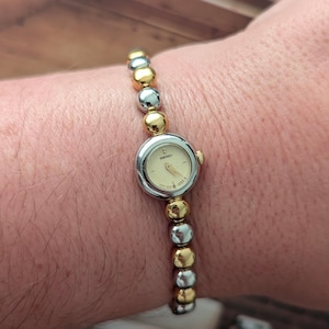 May include: A Seiko wristwatch featuring a round, cream-colored analog dial with gold-tone hands and markers. The watch case is silver-tone, and the bracelet is composed of alternating polished gold-tone and silver-tone spherical metal beads.