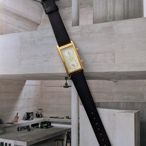 Puede incluir: Un reloj rectangular dorado con una correa de cuero negro. La esfera del reloj tiene dos esferas circulares con fondos blancos y n&uacute;meros negros. El reloj est&aacute; etiquetado como "KOLON".