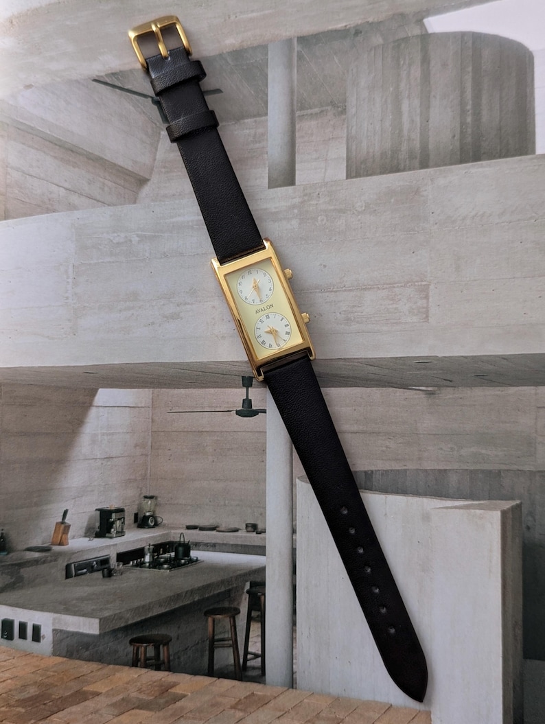 Puede incluir: Un reloj rectangular dorado con una correa de cuero negro. La esfera del reloj tiene dos esferas circulares con fondos blancos y n&uacute;meros negros. El reloj est&aacute; etiquetado como "KOLON".