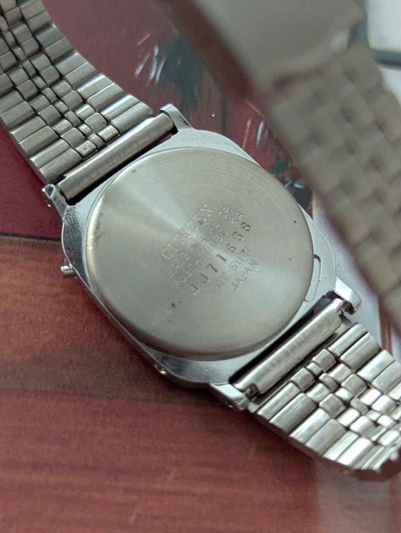May include: A silver-toned Citizen watch with a rectangular face and a metal band. The watch back displays the Citizen brand name and model information. The band is a flexible, woven style.