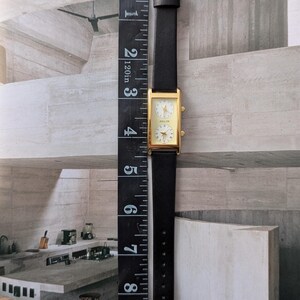 Puede incluir: Un reloj rectangular dorado con una correa de cuero negro. La esfera del reloj tiene dos esferas circulares con caras blancas y agujas doradas. El reloj mide aproximadamente 10 cm de largo, medido con una regla negra.