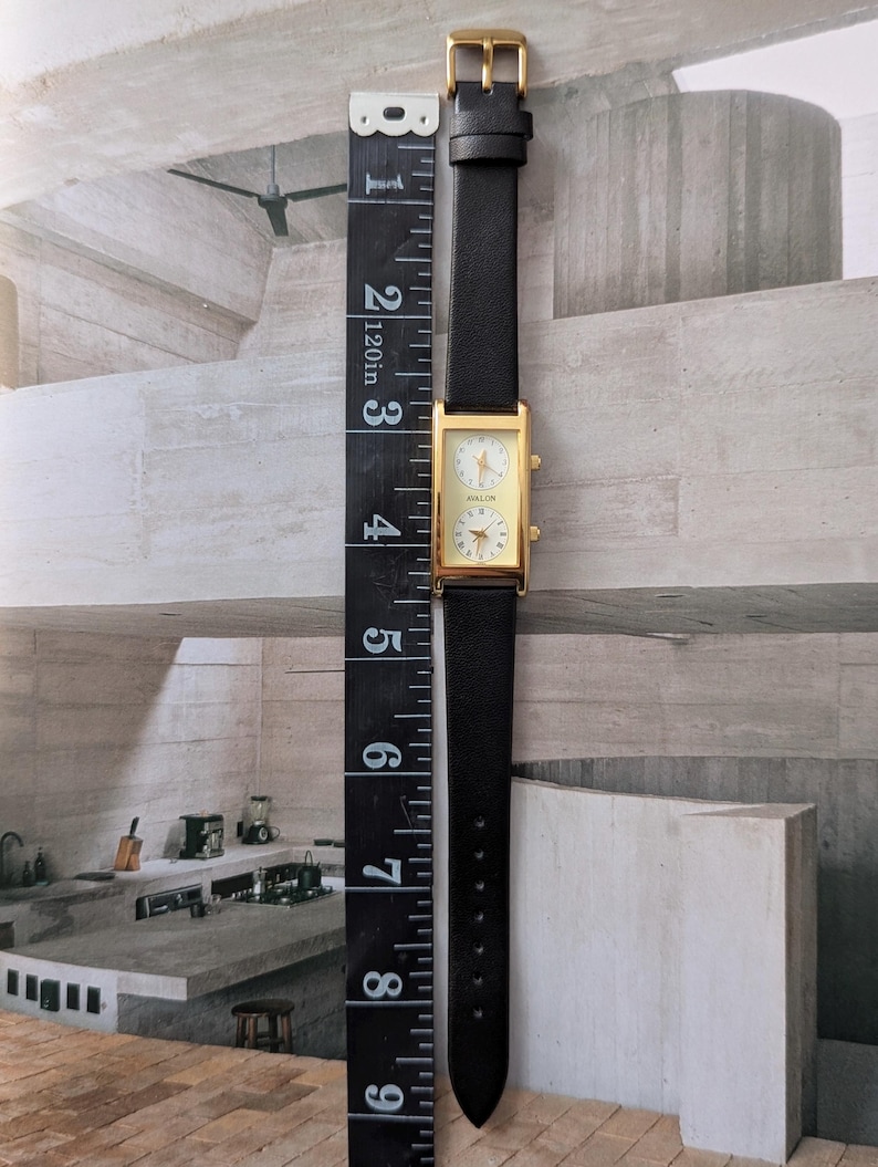 Puede incluir: Un reloj rectangular dorado con una correa de cuero negro. La esfera del reloj tiene dos esferas circulares con caras blancas y agujas doradas. El reloj mide aproximadamente 10 cm de largo, medido con una regla negra.