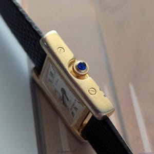 Peut inclure: Une montre-bracelet rectangulaire dor&eacute;e avec un cadran blanc, des aiguilles noires et des chiffres romains. La montre a un bracelet en cuir noir et une pierre pr&eacute;cieuse bleue sur la couronne. Design classique.