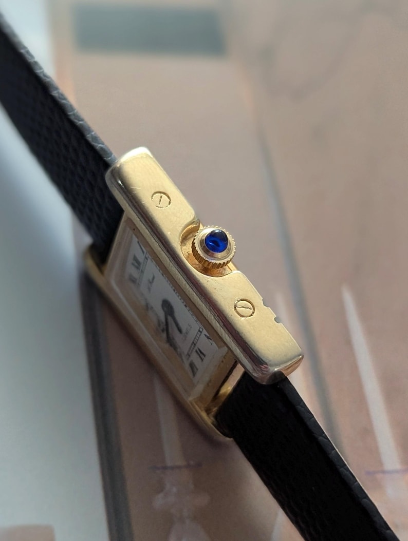 Peut inclure: Une montre-bracelet rectangulaire dor&eacute;e avec un cadran blanc, des aiguilles noires et des chiffres romains. La montre a un bracelet en cuir noir et une pierre pr&eacute;cieuse bleue sur la couronne. Design classique.