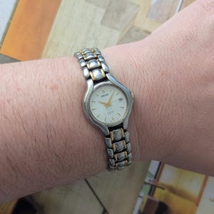 Puede incluir: Un reloj Seiko plateado y dorado con esfera blanca se lleva en la muñeca. El reloj tiene una esfera rectangular con agujas y marcadores dorados. La correa es una combinación de eslabones plateados y dorados.