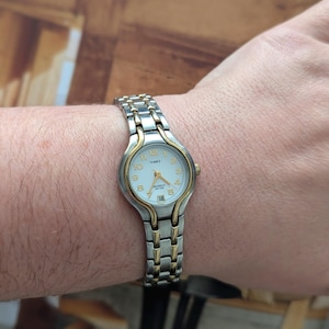 May include: A Timex watch with a silver and gold-tone metal band is shown on a wrist. The watch face is white with gold-tone hands and numerals. The date is displayed at the 6 o'clock position.
