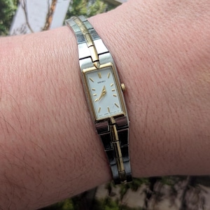 Puede incluir: Un reloj Seiko rectangular con una correa plateada y dorada. La esfera del reloj es blanca con agujas y marcadores dorados. El reloj se lleva en la muñeca.