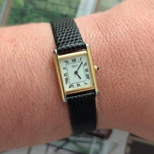 May include: A rectangular gold-tone Seiko Quartz wristwatch featuring a white dial with black Roman numeral hour markers and black hands. The watch has a textured black leather strap with a small crown on the right side of the case.