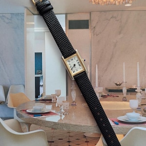 Peut inclure: Une montre-bracelet rectangulaire dor&eacute;e avec un cadran blanc et des chiffres romains noirs. La montre a un bracelet en cuir noir avec une boucle dor&eacute;e. La montre est pr&eacute;sent&eacute;e sur un fond de salle &agrave; manger.