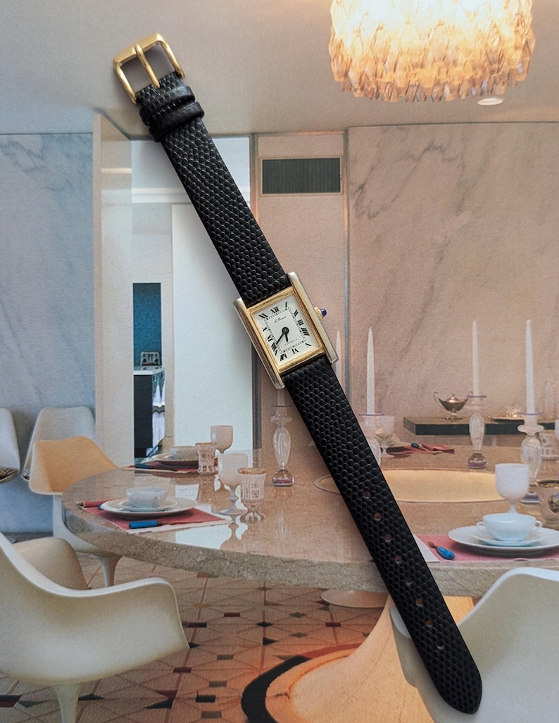 Peut inclure: Une montre-bracelet rectangulaire dor&eacute;e avec un cadran blanc et des chiffres romains noirs. La montre a un bracelet en cuir noir avec une boucle dor&eacute;e. La montre est pr&eacute;sent&eacute;e sur un fond de salle &agrave; manger.