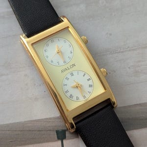 Puede incluir: Reloj rectangular dorado con dos esferas separadas, con esferas blancas y n&uacute;meros romanos negros. El reloj tiene una correa de cuero negro y la palabra "AVALON" est&aacute; impresa en la esfera.