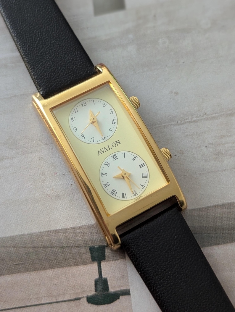 Puede incluir: Reloj rectangular dorado con dos esferas separadas, con esferas blancas y n&uacute;meros romanos negros. El reloj tiene una correa de cuero negro y la palabra "AVALON" est&aacute; impresa en la esfera.