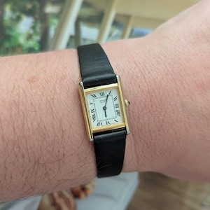 May include: A rectangular gold-toned wristwatch with a white face and black Roman numerals. The watch has a black leather strap. The watch face reads "SEIKO" and has gold-toned hands. The watch is on a wrist.