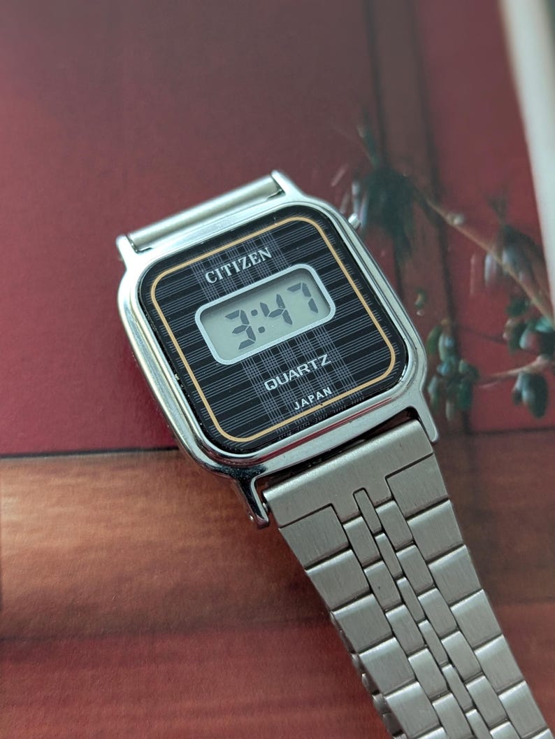 May include: A silver Citizen Quartz watch displaying the time 3:47. The watch has a black face with a grid pattern and gold trim. The watchband is made of silver metal links.