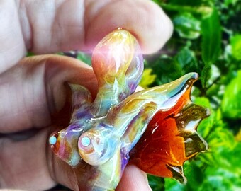 May include: A colorful, abstract glass sculpture with a flowing, organic form. The piece features a blend of rainbow hues, including orange, yellow, blue, and purple. Small, light blue accents are visible. The sculpture is held in a hand against a blurred green background.