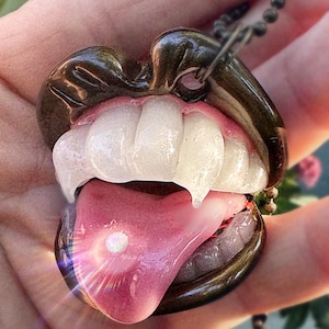 May include: A unique pendant necklace featuring a stylized mouth with fangs and a tongue. The lips are dark brown, the teeth are white, and the tongue is pink. The pendant is attached to a dark chain.