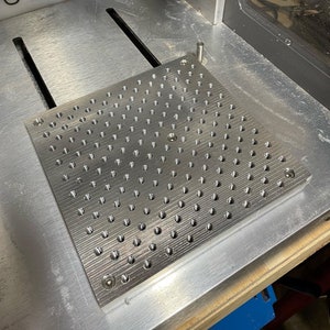 May include: A close-up of a silver metal plate with a grid of evenly spaced holes. The plate is sitting on a grey surface with the word "OMAD" visible in the background.