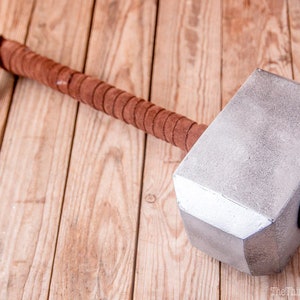 Puede incluir: Un martillo de color plateado con un mango de cuero marrón. El martillo tiene la forma del martillo de Thor, Mjolnir, con una cabeza redonda y un mango corto.