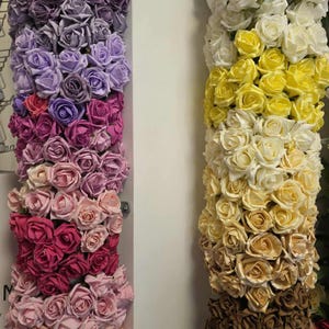 May include: A display of artificial roses in various colours, including purple, pink, and yellow. The roses are arranged in vertical columns, showcasing a gradient of shades from light to dark. The image is well-lit, highlighting the texture and detail of the flowers.