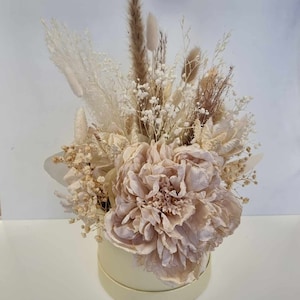 May include: A dried flower arrangement in a cream-coloured box. The arrangement features a large, blush-coloured peony, white baby's breath, and dried grasses.