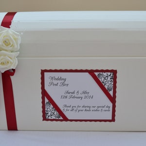 May include: Cream wedding post box with a curved lid, decorated with a red ribbon and white roses. A label displays the text "Wedding Post Box" and the names "Sarah & Alex". The box is designed for collecting wedding cards.