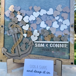 May include: A wooden guest book with a blue background and an anchor design. The text "Sam & Connie Anchored in Love" is written on the sign. The sign is surrounded by a wooden frame and has a glass front. There are small wooden hearts inside the frame. A sign below the guest book reads "Sign a shape and drop it in".