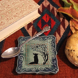 May include: A still life featuring a ceramic spoon rest with a black silhouette design, a silver spoon, a bagel, and a book. The spoon rest is square with a decorative border. The book has a patterned cover.
