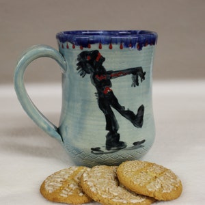 May include: A light blue ceramic mug with a zombie illustration. The mug has a blue handle and a dark blue rim with red drips. Three cookies with white icing are in front of the mug.