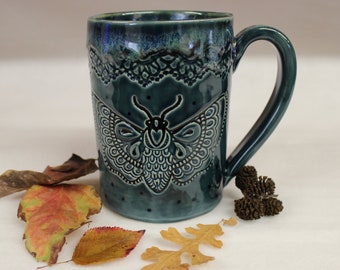 May include: A teal ceramic mug with a large handle, featuring an embossed moth design. The mug has a decorative lace pattern near the rim and is surrounded by fallen leaves, creating a seasonal aesthetic.