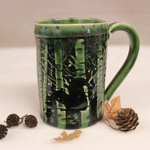 May include: A green ceramic mug with a forest scene design. The mug features a dark green forest scene with birch trees and a black silhouette. The handle and rim are a lighter shade of green. Decorative leaves and pine cones are placed near the mug.