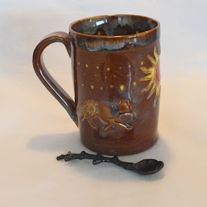 May include: A brown ceramic mug with a handle, featuring a sun and wolf design. The mug has a dark brown glaze with yellow and red accents. A black spoon with a twig-like handle rests beside the mug.