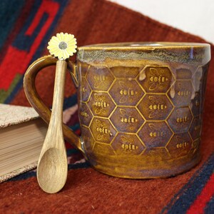 May include: A ceramic mug with a honeycomb pattern and bee designs, paired with a wooden spoon topped with a yellow flower. The mug has a brown and purple glaze. A book rests beside the mug on a red and blue patterned textile.