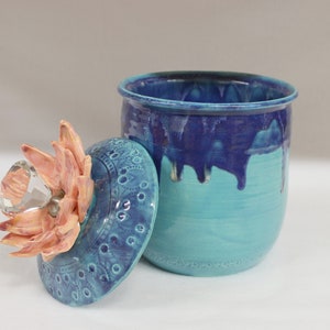 May include: A handcrafted ceramic jar with a lid. The jar is turquoise with a dark blue top that drips down the sides. The lid is turquoise with a floral design and a crystal knob. The flower is peach-colored.
