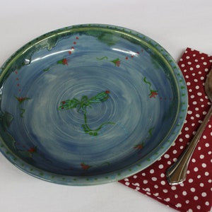 May include: A handmade ceramic serving bowl with a blue interior and green rim, decorated with a dragonfly and floral accents. A silver serving utensil rests on a red polka dot napkin.