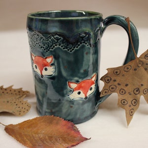 May include: A teal ceramic mug with a handle, featuring two orange and white fox faces. The top rim has a decorative textured pattern. The mug is approximately 4 inches tall.